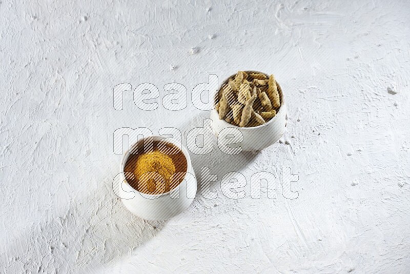 وعاءان فخاريان بيج ممتلئان ببودرة الكركم وأصابع الكركم المجففة على خلفية بيضاء