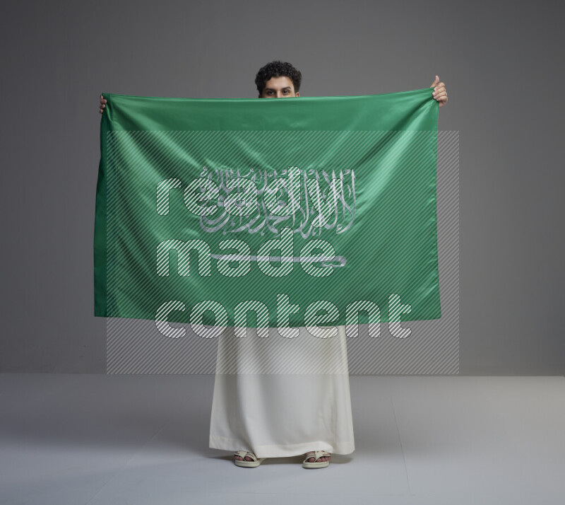 A Saudi man standing wearing thob holding big Saudi flag on gray background