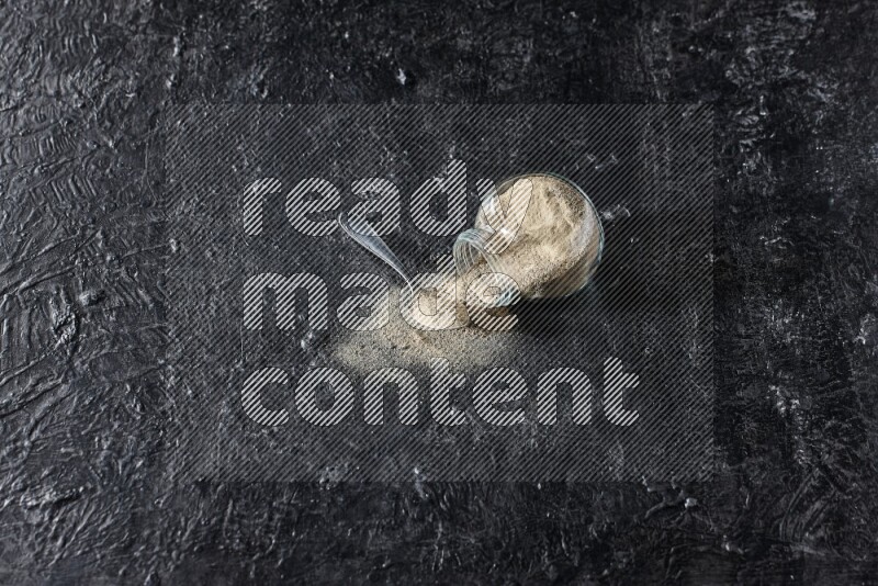 A flipped herbal glass jar and a metal spoon full of white pepper powder with spilled powder on textured black flooring
