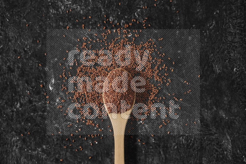 A wooden spoon full of garden cress seeds surrounded by the seeds on a textured black flooring