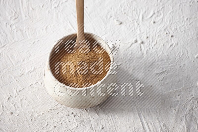 Ceramic beige bowl full of cinnamon powder with a wooden spoon on a textured white background