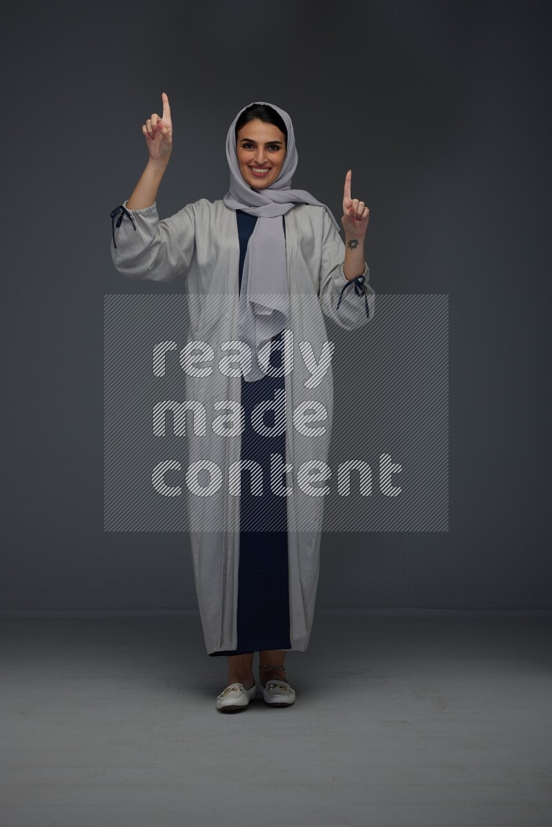 A Saudi woman wearing a light gray Abaya and head scarf standing and pointing in different directions eye level on a grey background