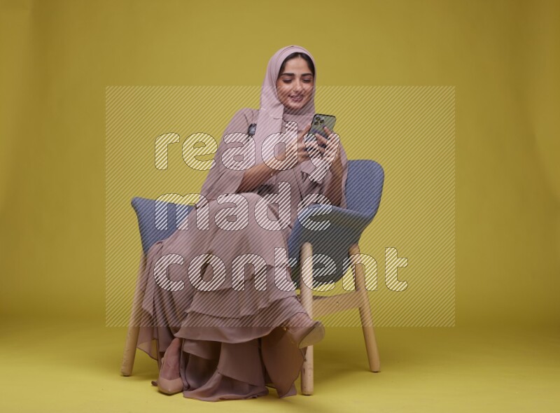 A Female Texting Sitting  on a Yellow Background wearing Brown Abaya with Hijab