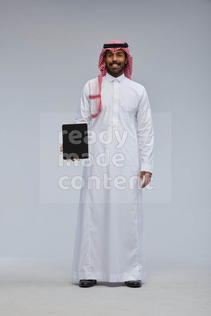 Saudi man Wearing Thob and shomag standing showing tablet to camera on Gray background