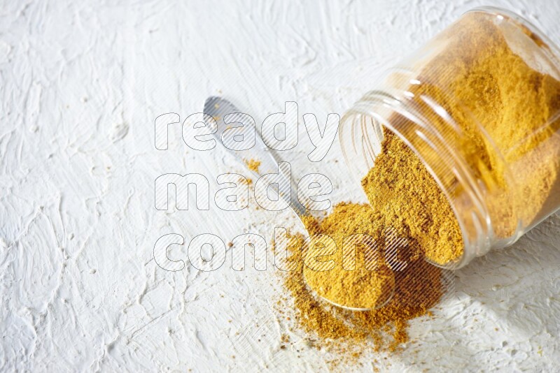 A flipped glass spice jar and a metal spoon full of turmeric powder and powder spilled out of it on textured white flooring