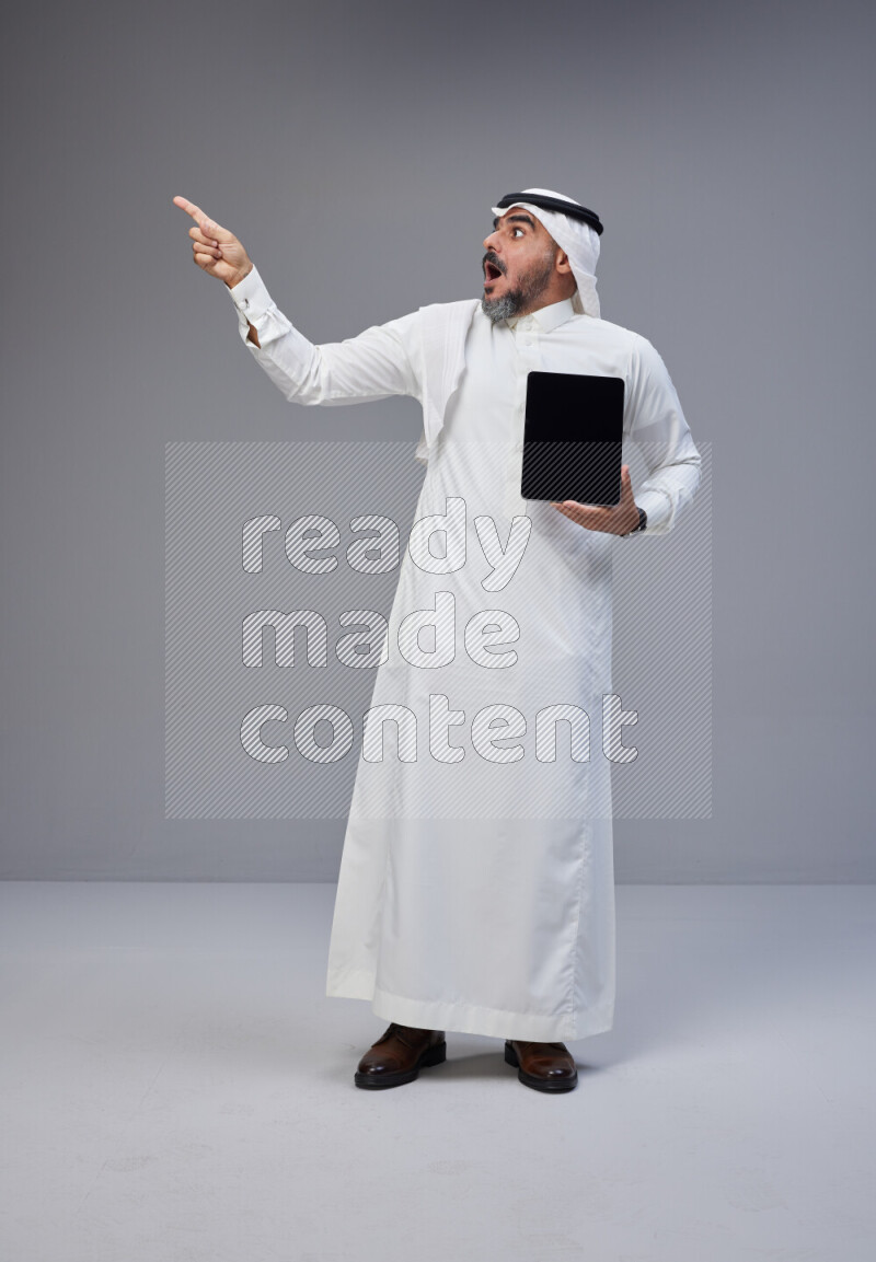 Saudi man Wearing Thob and white Shomag standing showing tablet to camera on Gray background
