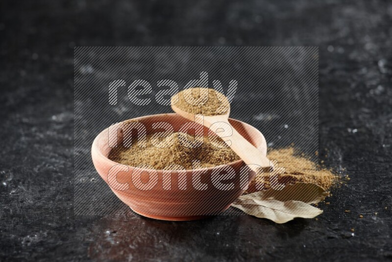 A wooden bowl and spoon full of cumin powder on a textured black flooring
