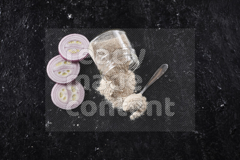 A glass jar full of onion powder flipped with some spilling powder on black background