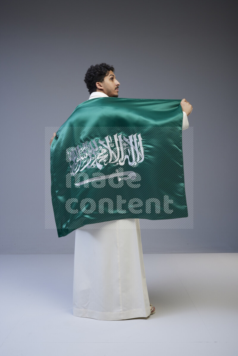 A Saudi man standing wearing thob holding big Saudi flag on gray background