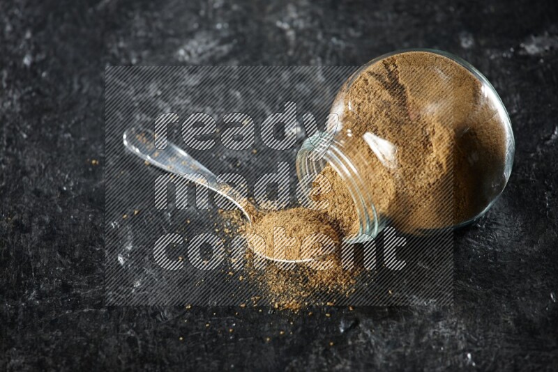 A flipped glass spice jar and a metal spoon full of cumin powder and powder spilled out on a textured black flooring