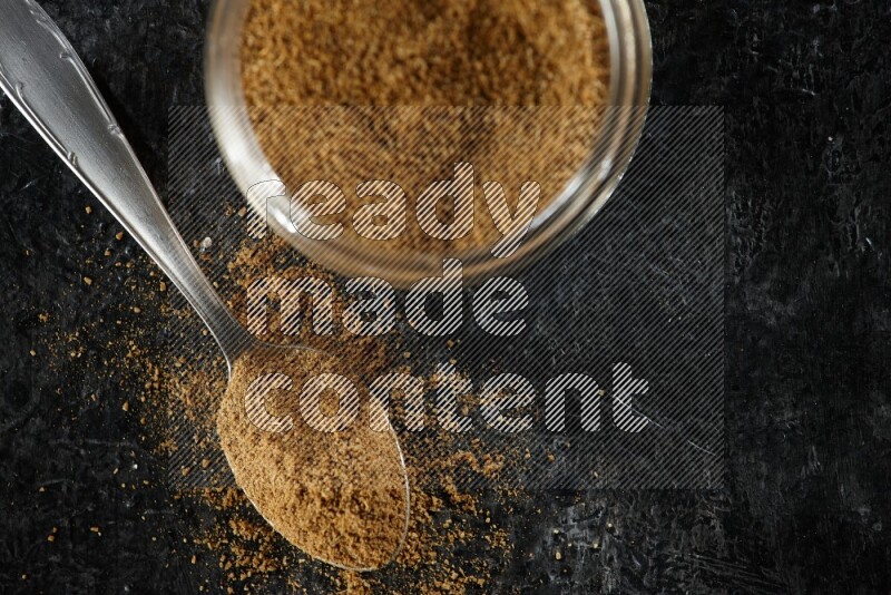 A glass jar and a metal spoon full of cumin powder on a textured black flooring