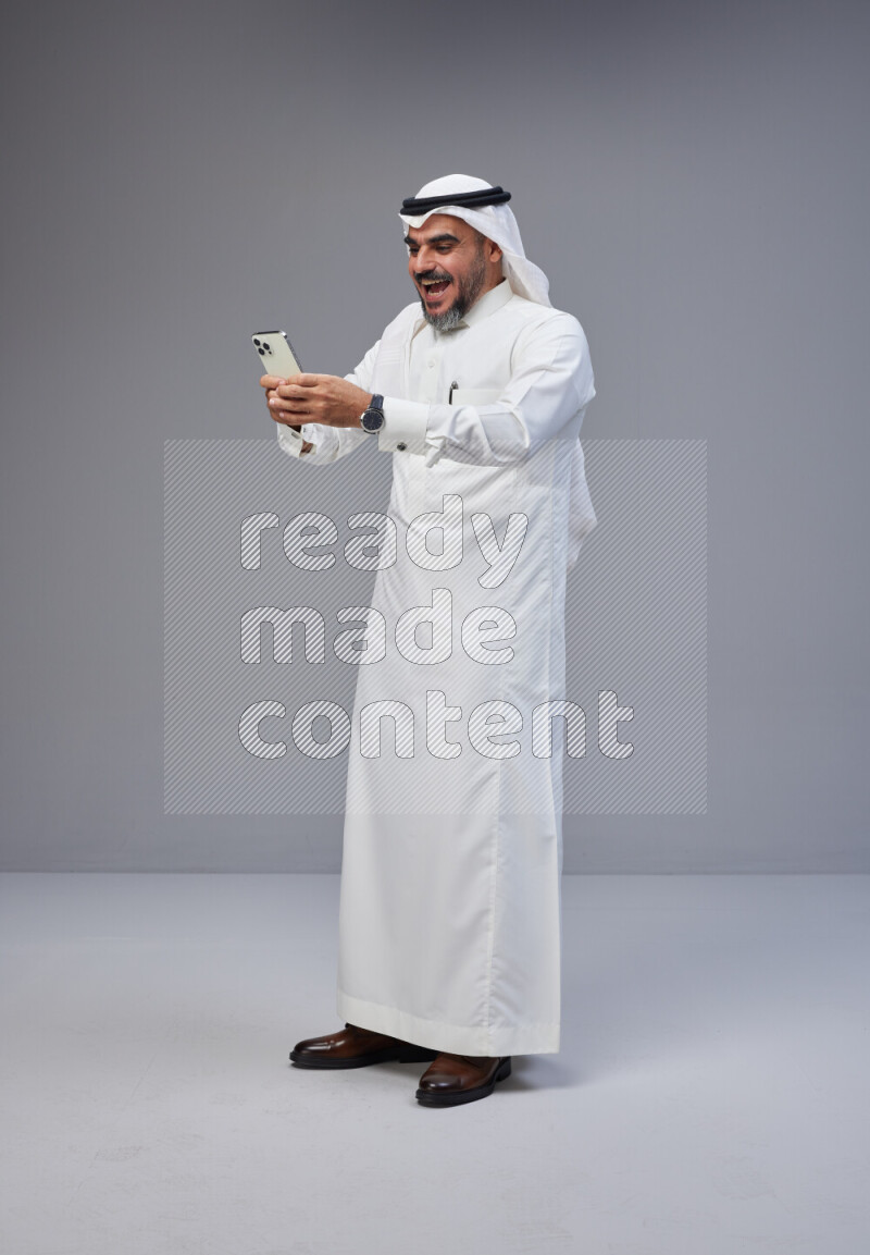 Saudi man Wearing Thob and white Shomag standing texting on phone on Gray background