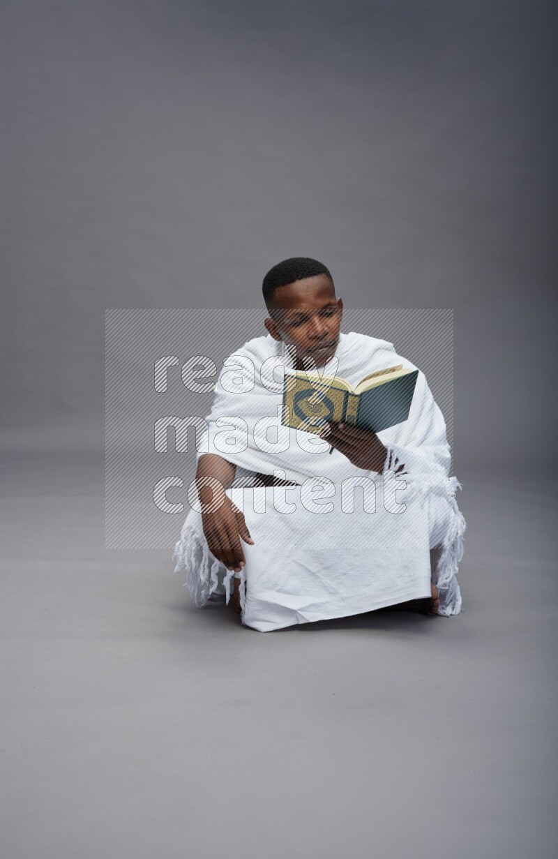 A man wearing Ehram sitting on floor reading quran on gray background