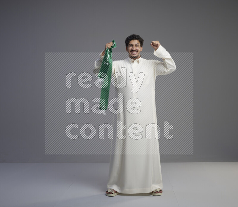 A Saudi man standing wearing thob holding Saudi flag scarf on gray background