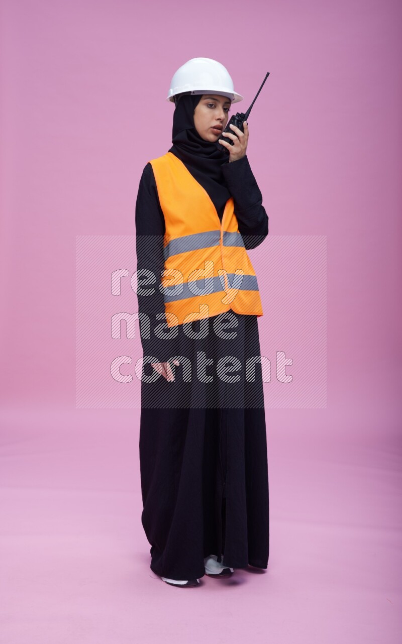 Saudi woman wearing Abaya with engineer vest and helmet standing holding walkie-talkie on pink background