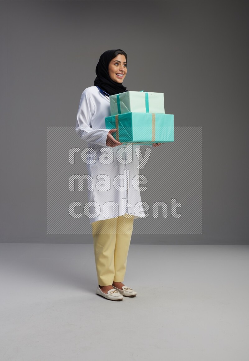 Saudi woman wearing lab coat with stethoscope standing holding gift box on Gray background