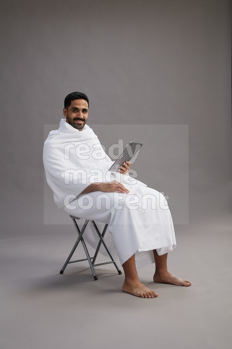 A man wearing Ehram sitting on chair holding a tablet on gray background