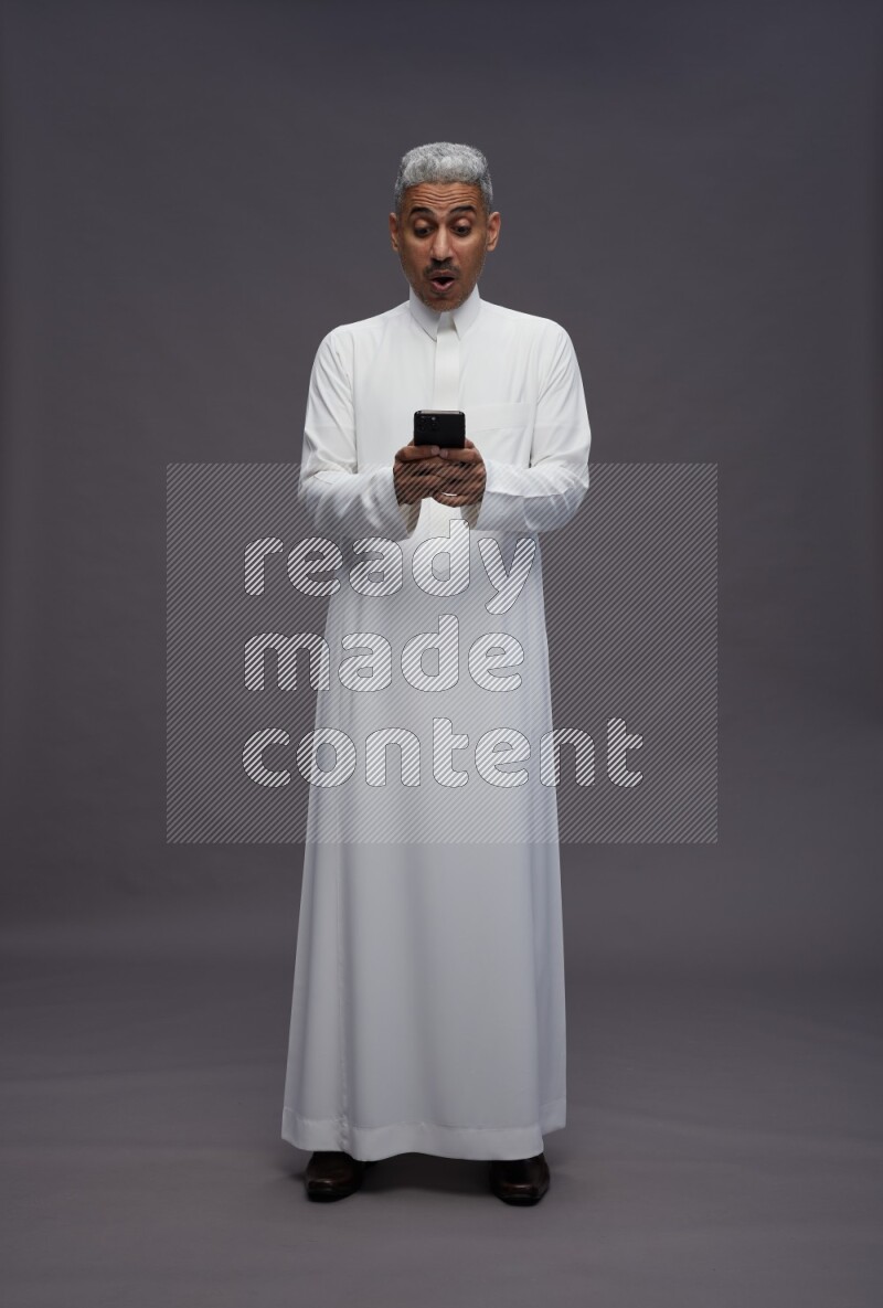 Saudi man wearing thob standing texting on phone on gray background