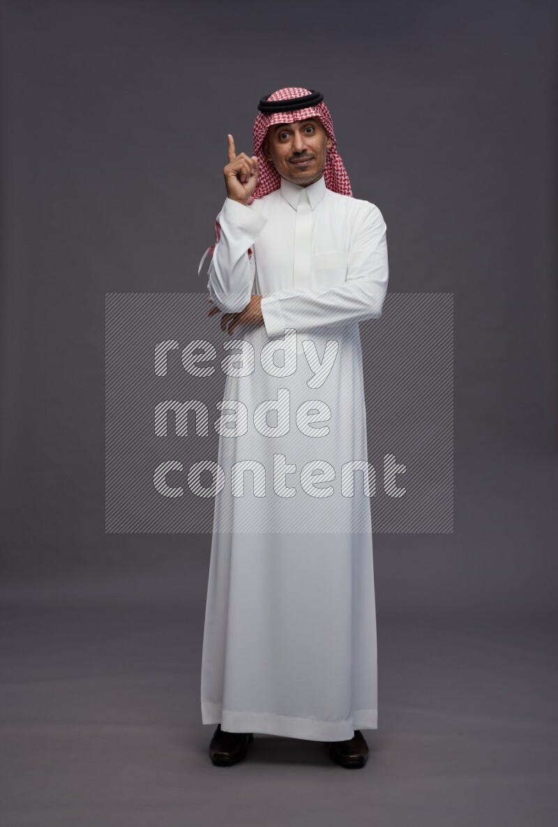 Saudi man wearing thob and shomag standing interacting with the camera on gray background