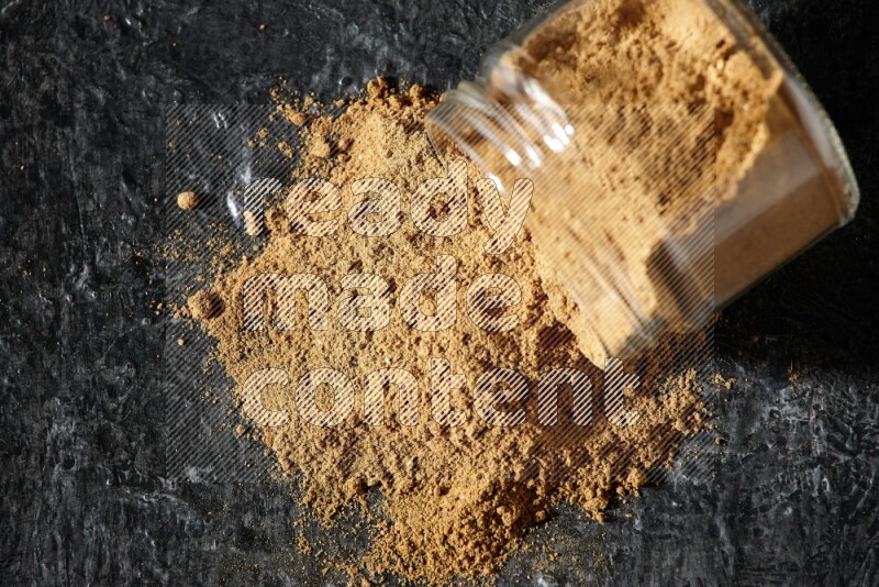 A flipped glass jar full of allspice powder and powder spilled out of it on a textured black flooring