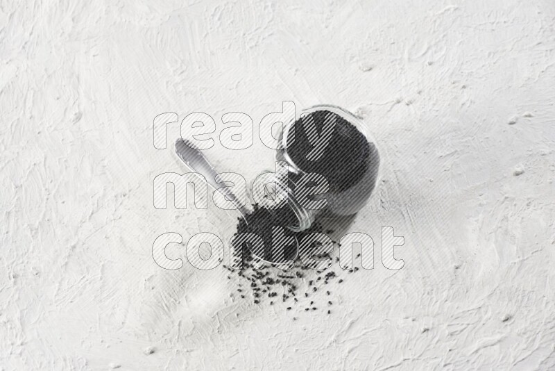 A glass spice jar and a metal spoon full of black seeds and the jar flipped and seeds spread on a textured white flooring