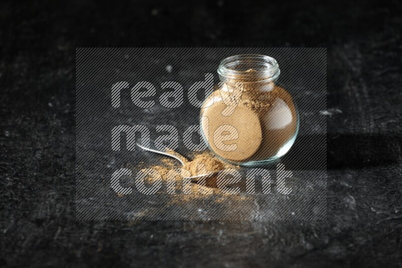 A glass spice jar and metal spoon full of allspice powder on a textured black flooring