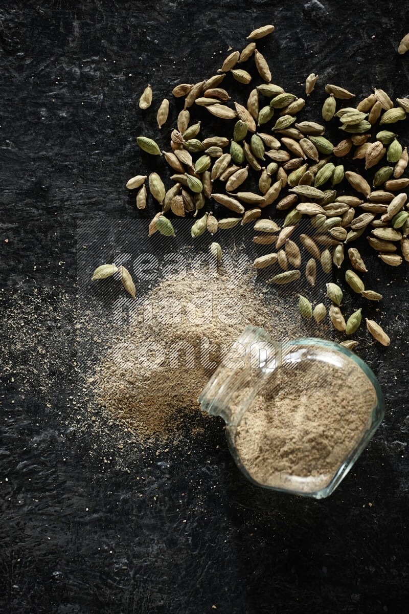 وعاء زجاجي للتوابل مقلوب وممتلئ ببودرة الهيل والبودرة تناثرت منه مع حبوب الهيل المتناثرة على خلفية سوداء