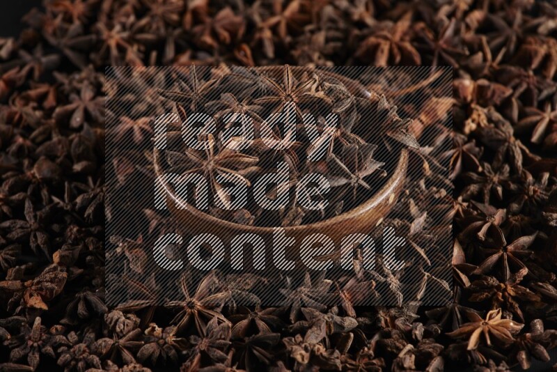 Star Anise in a wooden bowl and surrounded by more anise filling the frame on black flooring