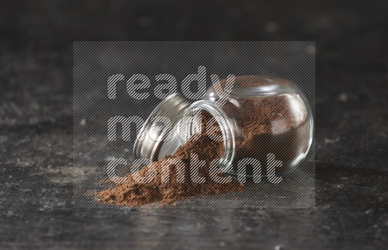 A flipped glass spice jar full of cloves powder and powder came out of it on textured black flooring