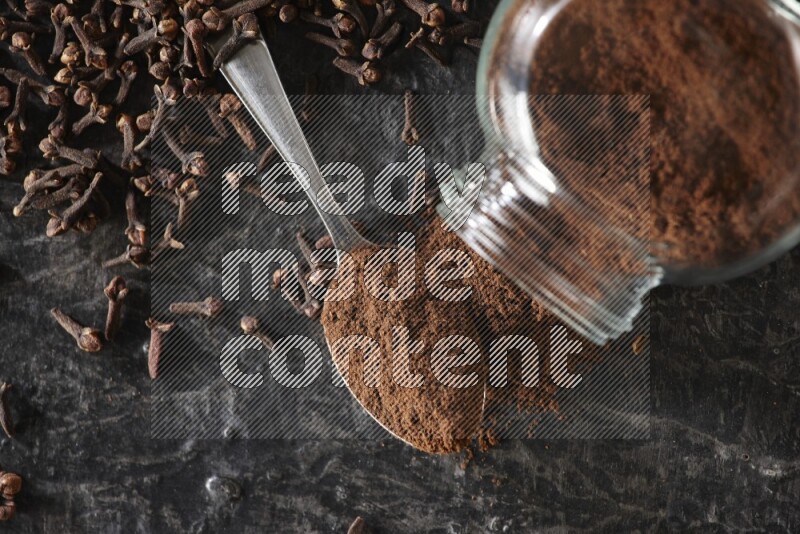 A flipped glass spice jar and a metal spoon full of cloves powder with cloves spread on textured black flooring