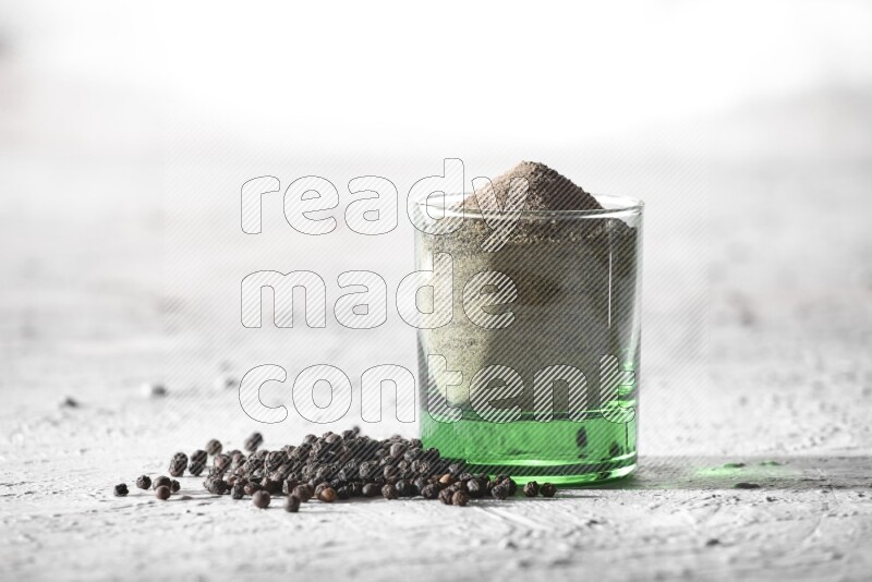 A glass cup full of black pepper powder and black pepper spread on a textured white flooring