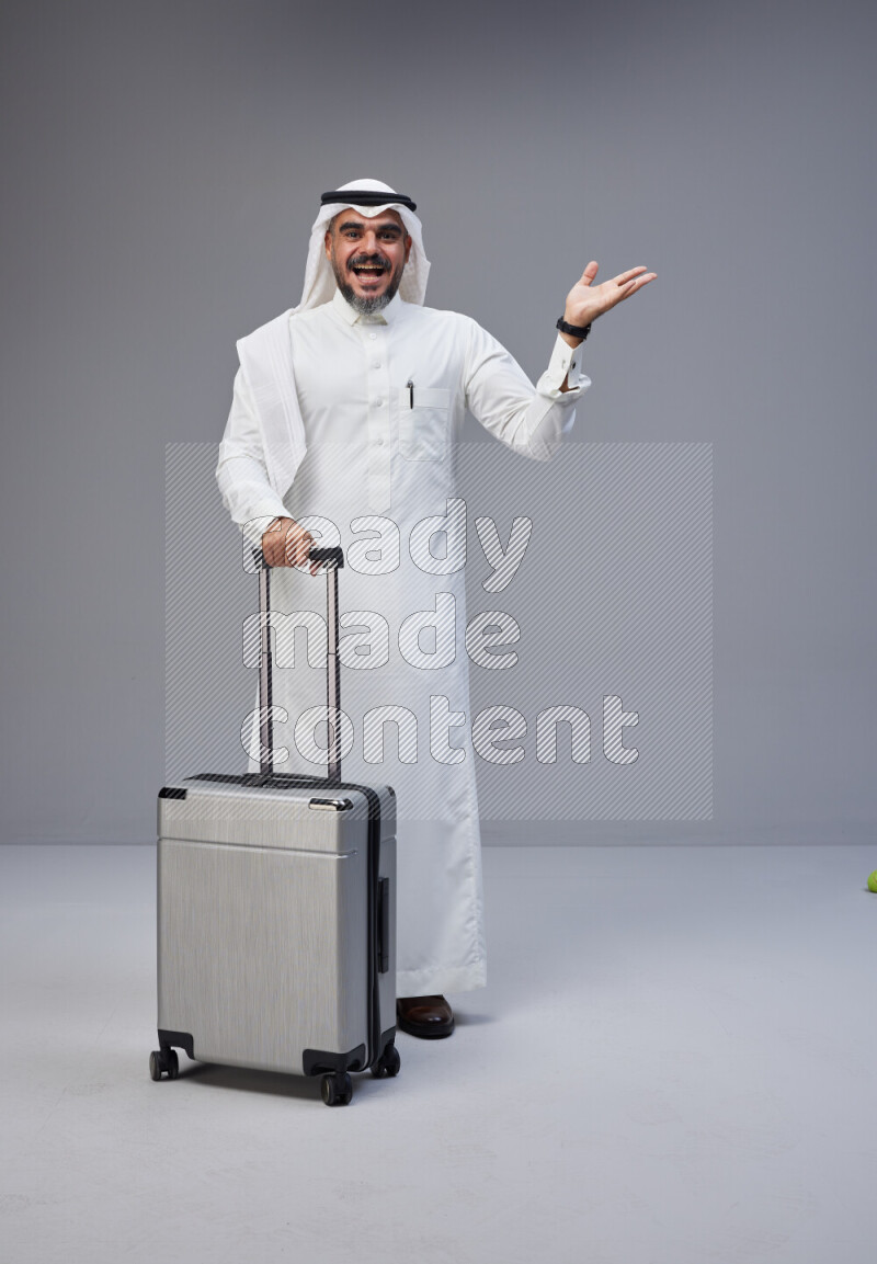 Saudi man wearing Thob and white Shomag standing holding Travel bag on Gray background