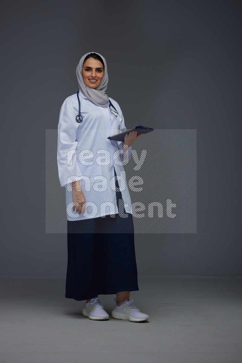 A Saudi doctor wearing a light gray head scarf standing and crossing her hands eye level on a grey background