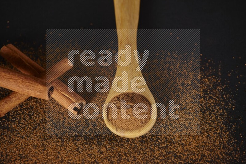 Cinnamon powder in a wooden spoon and cinnamon sticks beside it on black background