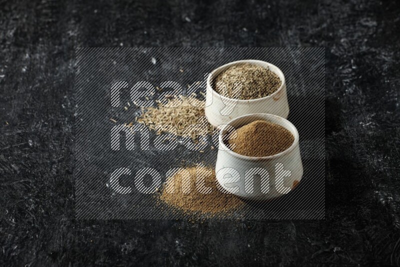 2 beige bowls, one full of cumin powder and the other full of seeds on a textured black flooring
