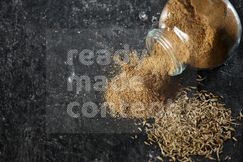 وعاء زجاجي مقلوب ممتلئ ببودرة الكمون مع بودرة متناثرة وحبوب الكمون على خلفية سوداء