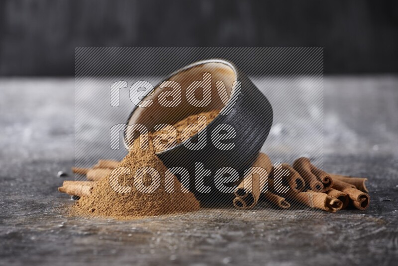 Black pottery bowl over filled with cinnamon powder and cinnamon sticks around the bowl on a textured black background