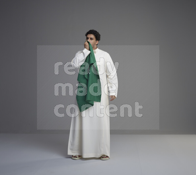 A Saudi man standing wearing thob kissing Saudi flag on gray background