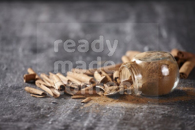 Herbal glass jar full cinnamon powder flipped and a metal spoon full of powder surrounded by cinnamon sticks on textured black background in different angles