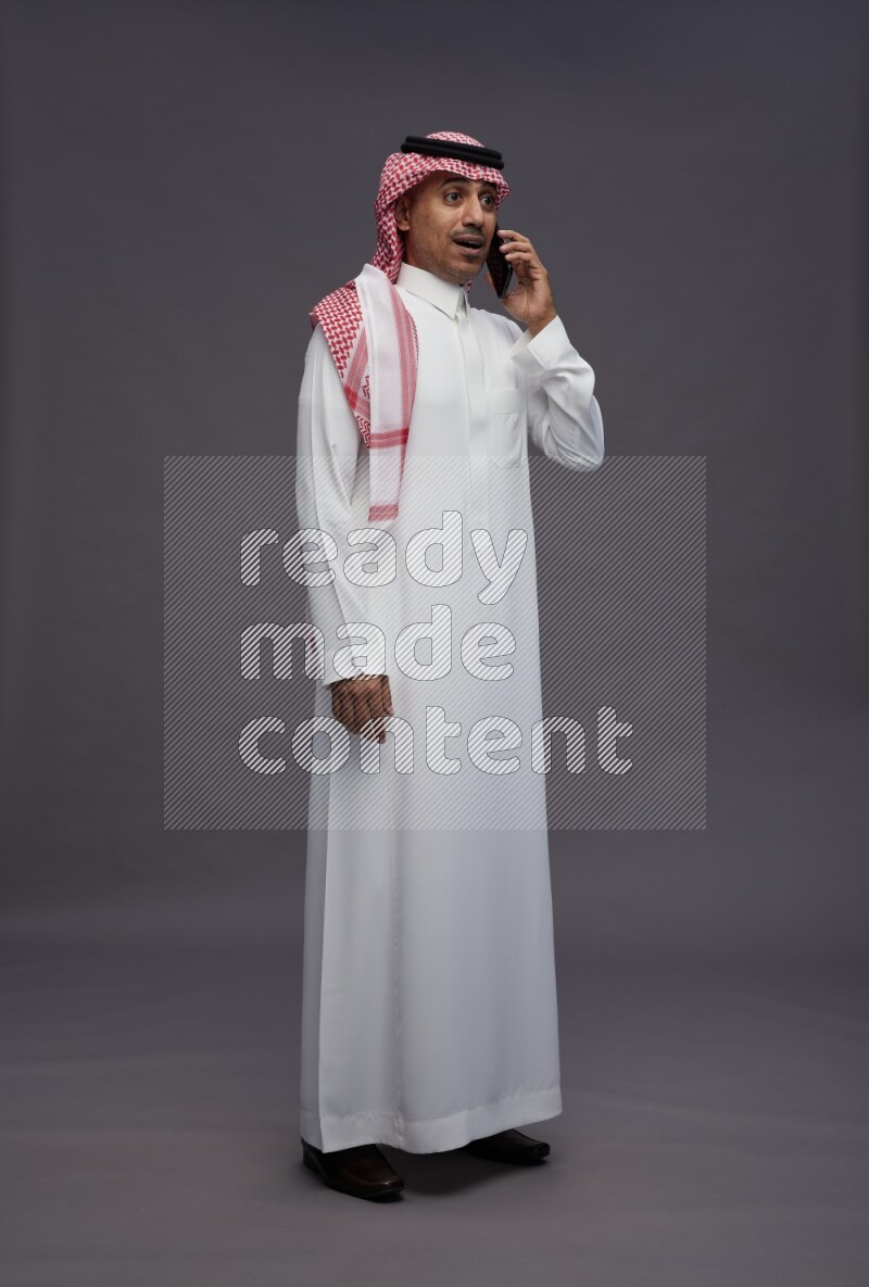 Saudi man wearing thob and shomag standing talking on phone on gray background