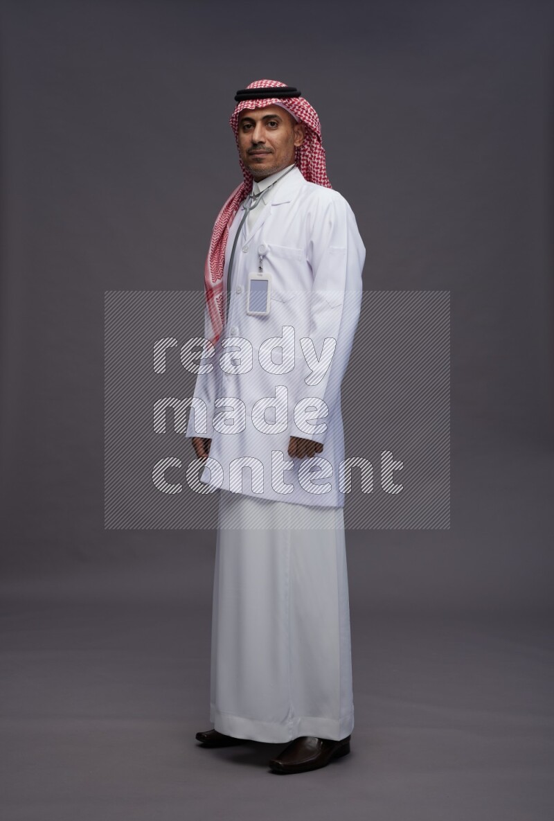 Saudi man wearing thob with lab coat and shomag with pocket employee badge with stethoscope standing interacting with the camera on gray background