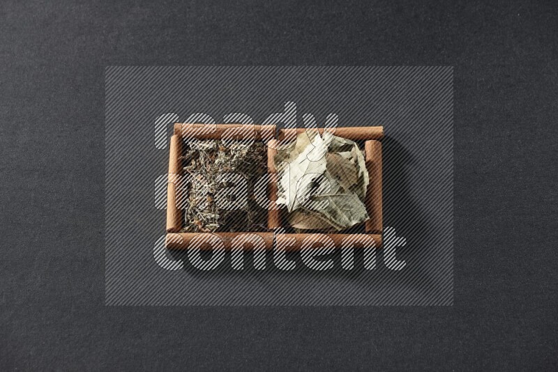 مربعان من أعواد القرفة ممتلئان بأوراق الغار والريحان المجفف علي خلفية سوداء