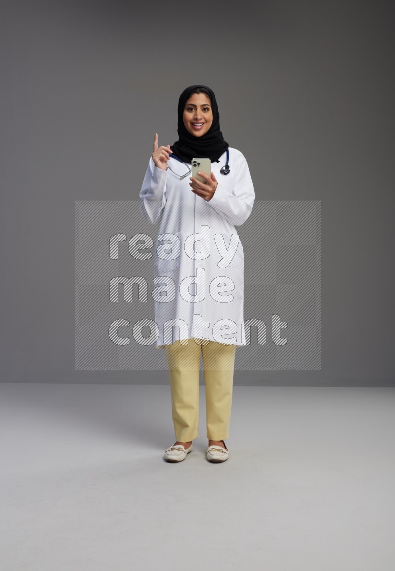Saudi woman wearing lab coat with stethoscope standing texting on phone on Gray background