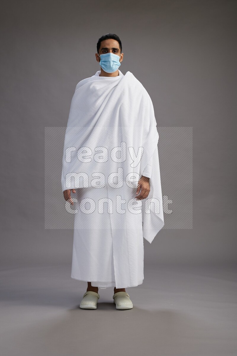 A man wearing Ehram with face mask Standing Interacting with the camera on gray background