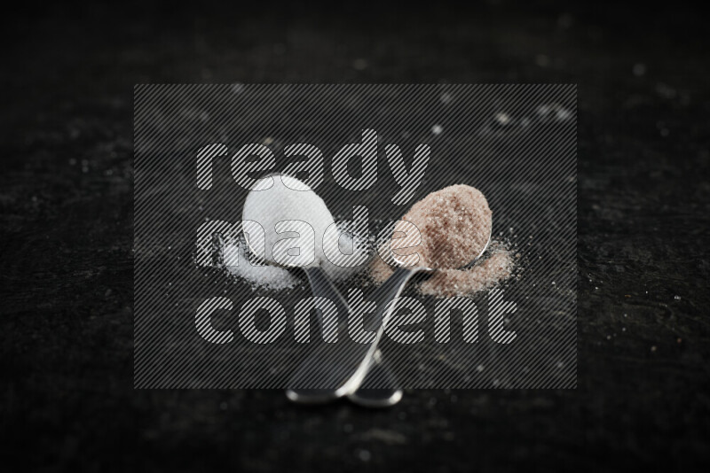 2 metal spoons filled with white salt and pink himalayan salt on black background
