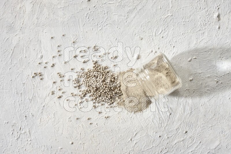 A flipped glass jar full of white pepper powder with spilled powder and white pepper beads on textured white flooring