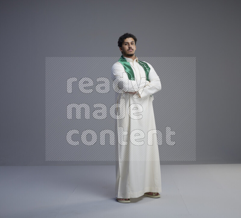 A Saudi man standing wearing thob and Saudi flag scarf on gray background