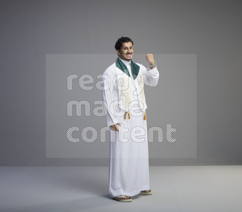 A Saudi man standing wearing thob and Saudi flag scarf with face painting on gray background