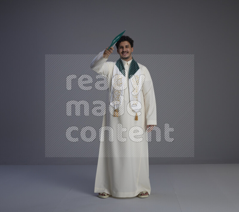A Saudi man standing wearing thob and saudi flag scarf and holding small saudi flag on gray background