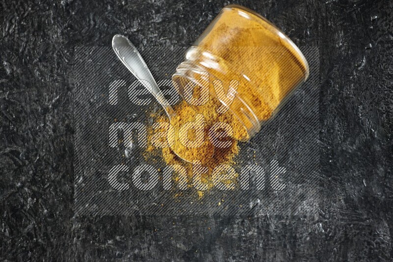 A flipped glass jar and a metal spoon full of turmeric powder and powder spilled out of it on textured black flooring